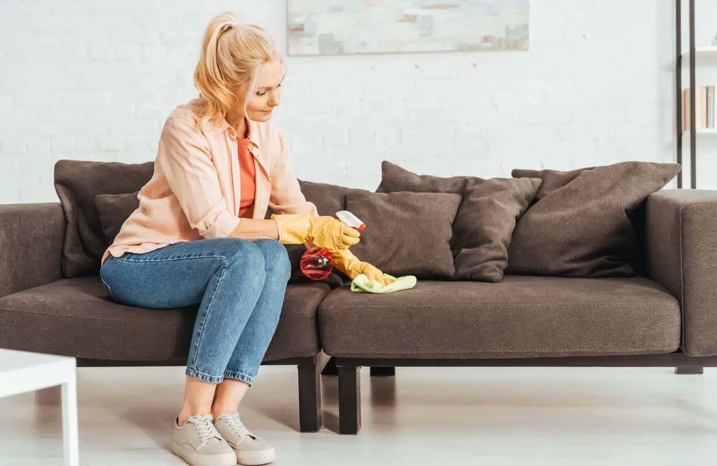 blonde woman cleaning brown couch cushions with a green microfibre cloth