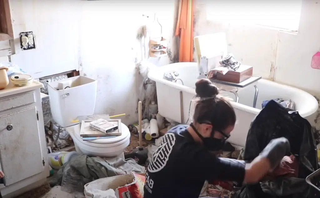 A bathroom full of rubbish and a woman wearing a face mask cleaning