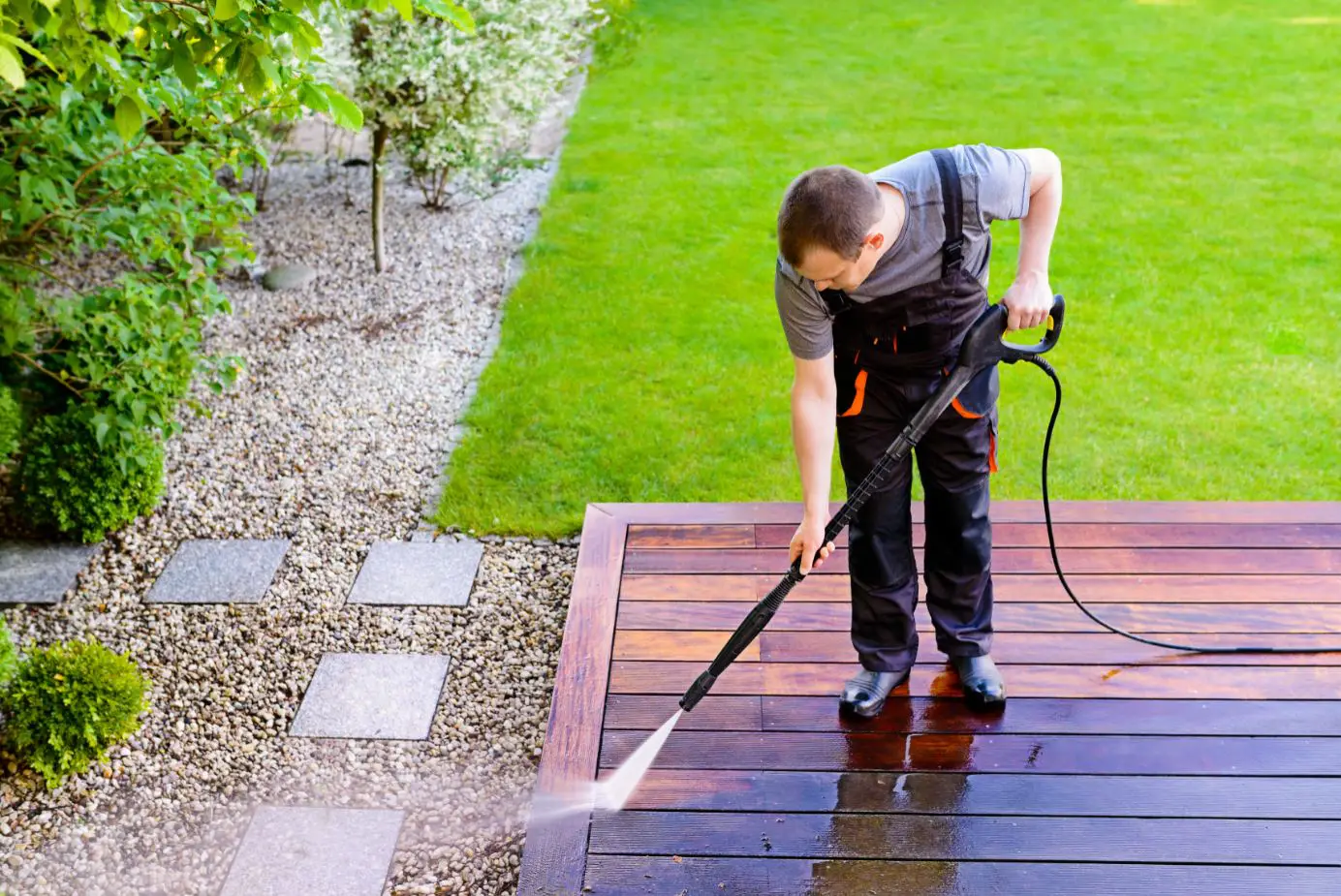 man in dark coloured overalls bending down to pressure clean the side of an outside patio