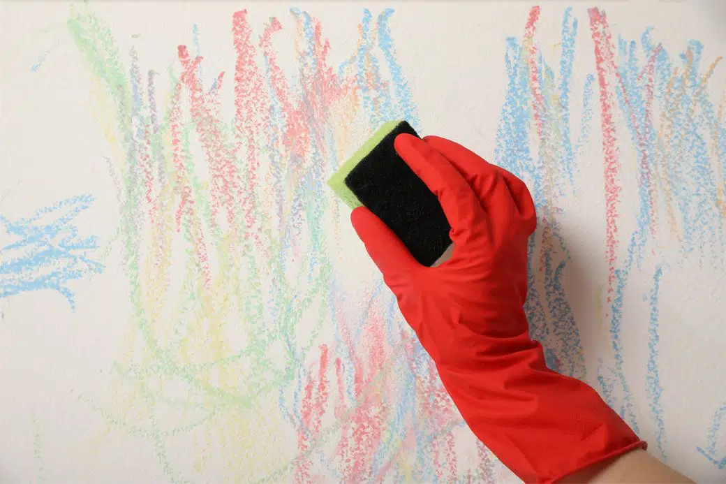 person wearing red gloves using a sponge to clean crayon off white wall