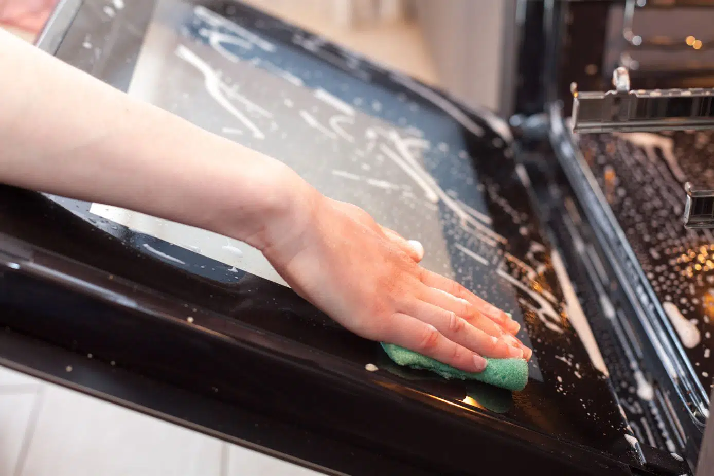 woman cleaning oven glass with a soapy green sponge